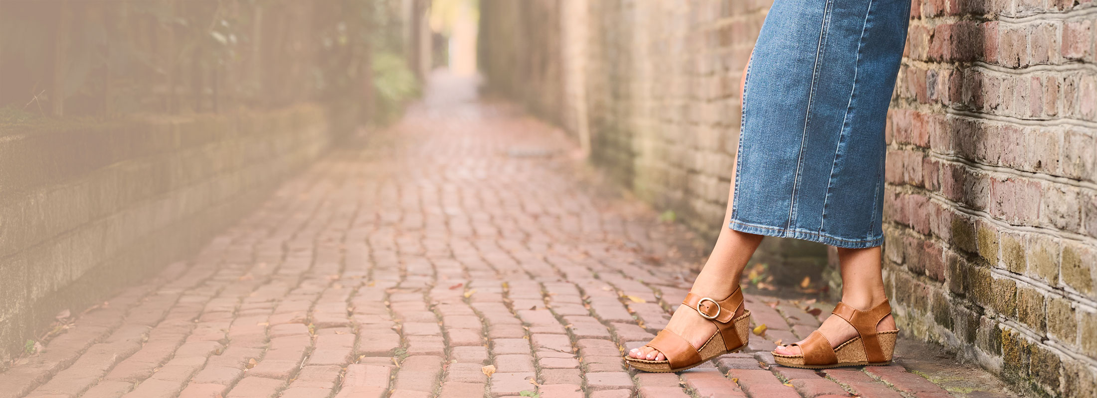 A woman standing against a brick wall wearing the Eldorah sandals in tan.