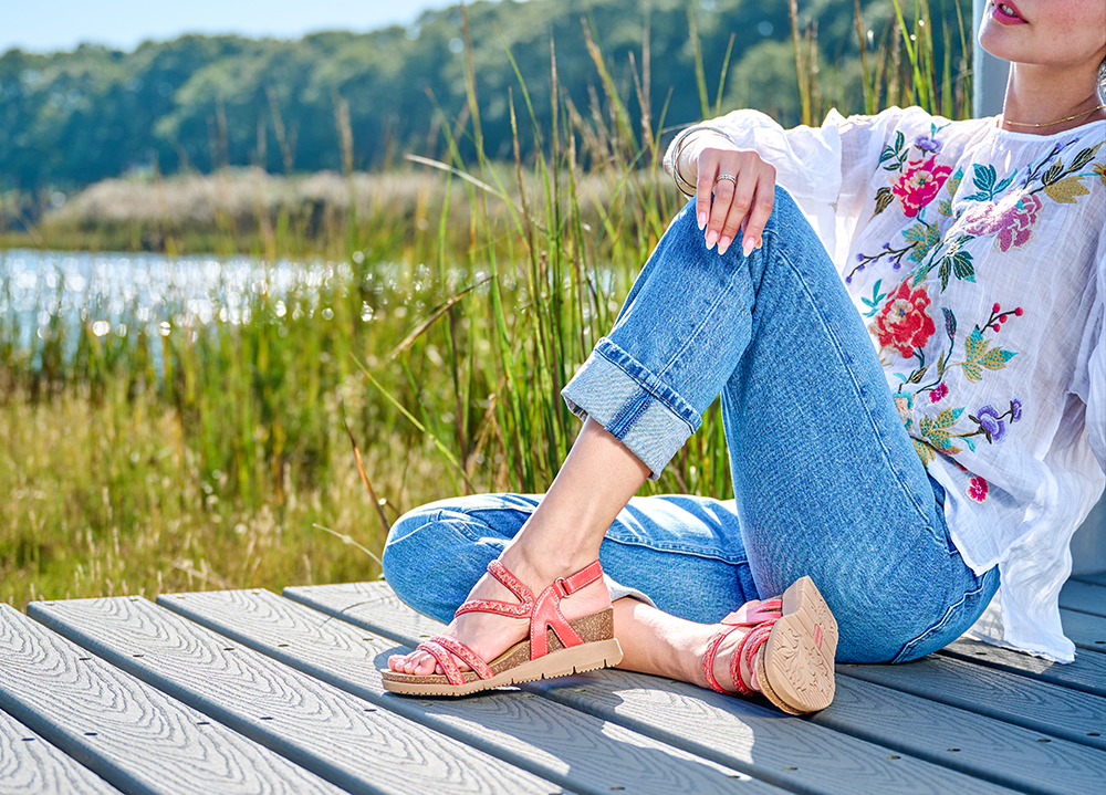 A woman sitting on a dock wearing Silvia sandal in Tomato Multi (red)