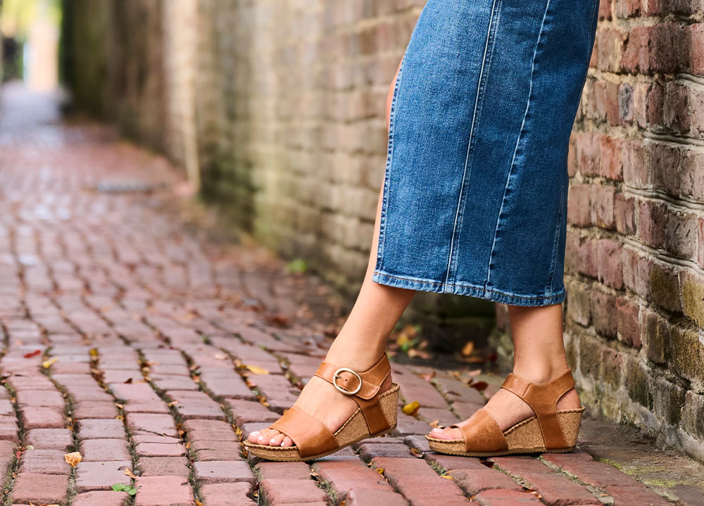 A woman standing against a brick wall wearing the Eldorah sandals in tan.